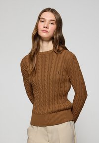 Brązowy sweter w splot kablowy z prążkowanym dołem i mankietami. Ma okrągły dekolt oraz subtelny detal z logo na piersi. Materiał wygląda na teksturowany.