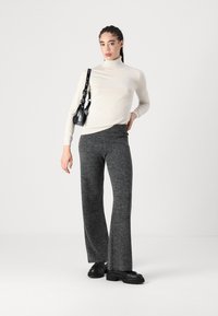 Pulover bej cu guler înalt, asociat cu pantaloni tricotati gri, cu croială largă. Modelul poartă pantofi negri și ține în mână o geantă mică neagră.