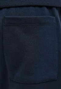 Close-up van donkerblauwe stof met een rechthoekig aangebracht zakje waarop zichtbare stiksels langs de randen te zien zijn.