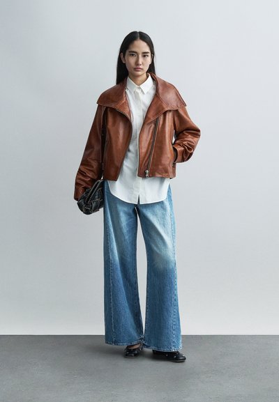 Καφέ δερμάτινο μπουφάν με φαρδιές πέτσες, λευκό πουκάμισο με κουμπιά και χαλαρό μπλε τζιν παντελόνι. Μαύρη τσάντα με λουρί αλυσίδα. Πλακέ παπούτσια.