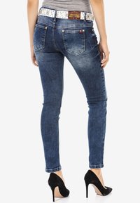 Jean skinny en denim de lavage bleu foncé avec une texture légèrement déchirée et cinq poches. Porté avec une ceinture blanche à clous et des escarpins noirs.