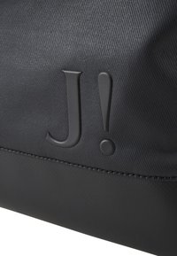 Črn platneni kovček z teksturirano površino, embosiranim logotipom "J!" in okroglim dodatkom. Oblikovanje je elegantno z trdno konstrukcijo.