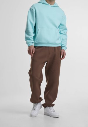 Person trägt hellblauen Kapuzenpullover, braune weite Hose und weiße Turnschuhe, steht vor einem einfachen hellen Hintergrund.