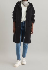 Czarna, dłuższa kurtka z kapturem, wykonana z miękkiego materiału, z talią na sznurki i zapięciem na zamek, w połączeniu z jasnoniebieskimi dżinsami i białymi trampkami.