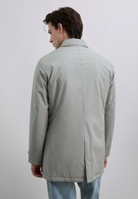 Joven de espaldas vistiendo una chaqueta de corte a la rodilla de color gris claro y jeans azul claro, frente a un fondo gris liso.