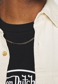 Camisa beige de botones sobre una camiseta negra de canalé con logo blanco, adornada con un delicado collar de cadena dorada, cuellos cortos y tela texturizada.
