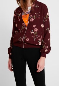 Veste bomber bordeaux avec motif floral, fermeture éclair et poignets côtelés. Associée à un t-shirt à motif graphique et des leggings noirs.