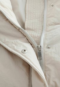 Pufjakke med en lys beige overflade, der har en glat tekstur, quiltet mønster, fremtrædende sølv lynlås og trykknapdetalje.