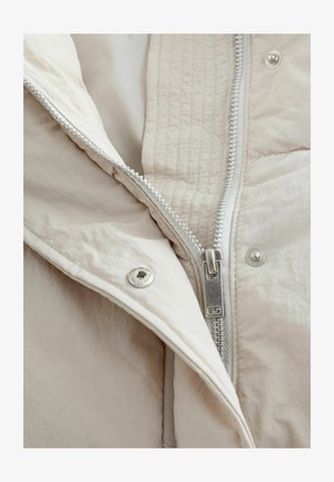 Puhumise jakk, millel on helebeež väline kiht, sile tekstuur, tikandiga muster, silmapaistev hõbedane tõmblukk ja nööbiga detail.