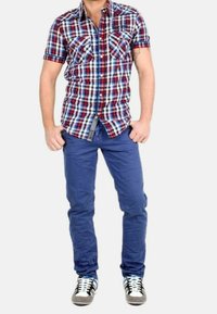 Camisa de cuadros en rojo, blanco y azul de manga corta con dos bolsillos en el pecho, combinada con pantalones ajustados azules y zapatillas con acentos en gris, blanco y negro.