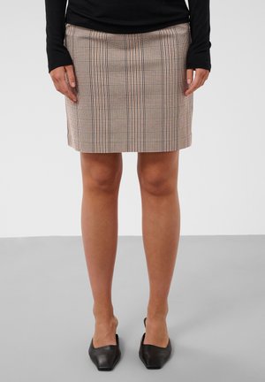 Vrouw draagt een beige geruite mini-rok, zwarte top met lange mouwen en zwarte slip-on schoenen met een vierkante neus, staand op een grijze vloer.