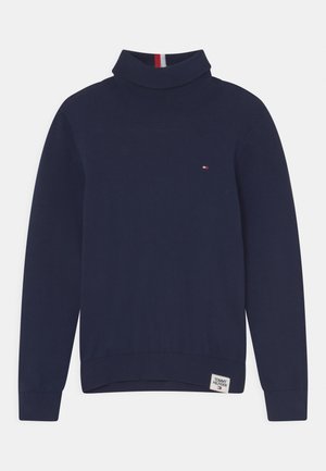 Pull à col roulé bleu marine à manches longues avec petit logo rouge, blanc et bleu sur la poitrine et étiquette Tommy Hilfiger à l'ourlet.