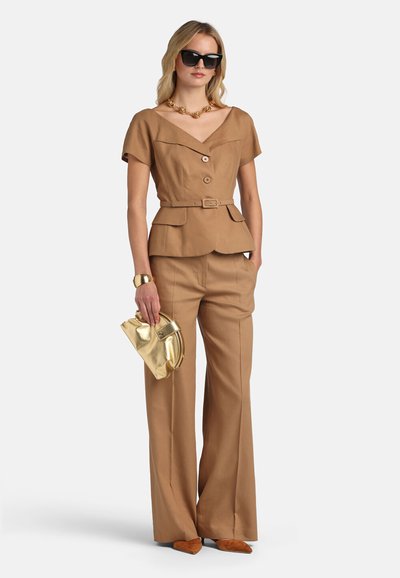 Vrouw met een tan blouse met korte mouwen en ceintuur, en wijde broekspijpen, houdt een gouden clutch vast, gecompleteerd met zwarte zonnebril en gouden sieraden.