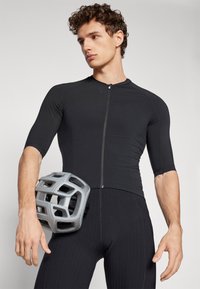 Maillot de cyclisme noir à manches courtes, avec fermeture éclair. Le modèle tient un casque gris avec des trous de ventilation. Design ajusté, texture lisse.