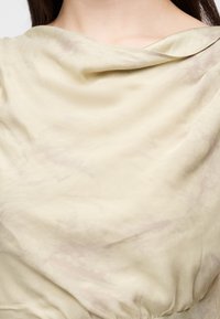 Gros plan sur une personne portant un haut en tissu drapé jaune pâle avec des détails froncés près de la taille, montrant le décolleté et les épaules.