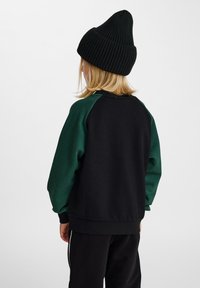 Svart och grön tröja med raglanärmar, mjuk textur, ribbade muddar och rundad halsringning. Stickad svart mössa på huvudet.