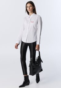 Mujer vestida con una camisa blanca a rayas, leggings de cuero negros y botas negras hasta el tobillo, sosteniendo un gran bolso negro con una correa de hombro.