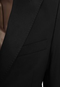 Zwarte colbert gemaakt van gladde stof, met een puntige revers en een klein paspelzakje aan de linkerkant. Schone stikdetails zijn zichtbaar.