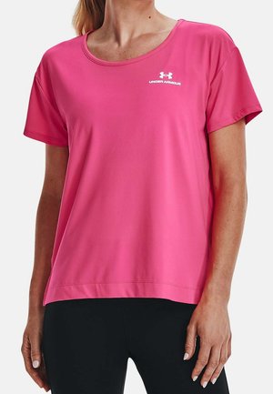Kobieta ubrana w jaskraworóżowy krótki rękaw Under Armour i czarne spodnie, stoi z rozluźnionymi ramionami przy bokach.