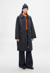 Μαύρο παλτό με καπιτονέ σχέδιο, γιακά, εμπρόσθιες τσέπες και χρυσές μεταλλικές λεπτομέρειες· φορεμένο πάνω από πορτοκαλί πουλόβερ και συνδυασμένο με τζιν φαρδιάς γραμμής και μπότες.