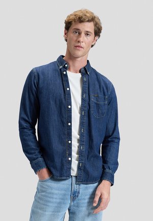 Jeune homme portant une chemise en jean bleu foncé déboutonnée sur un T-shirt blanc et un jean bleu clair, debout avec une main dans la poche.