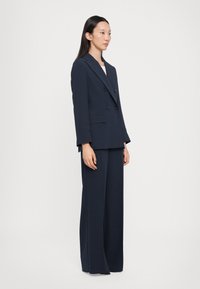 Námornícky modrý dvojradový blazer s chlopňovými klopami a vreckami na klapku, skombinovaný so širokými nohavicami. Hladká látka, strih na mieru.