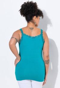 Camiseta de tirantes en color verde azulado con borde de encaje en el escote y en las tirantes. Tela suave y ajustada con una textura lisa. Pantalones blancos parcialmente visibles.