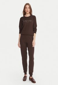 Felpa marrone e pantaloni jogger abbinati in tessuto morbido con polsini a costine. Un logo con strass è presente sulla parte anteriore della felpa.