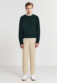 Felpa a maglia verde scuro con scollatura rotonda, abbinata a pantaloni beige su misura. Sneakers bianche completano l'outfit.