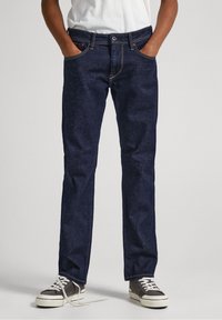 Jeans de denim azul oscuro con un corte recto, bolsillos frontales y costuras en contraste. Combinados con zapatillas grises y blancas.