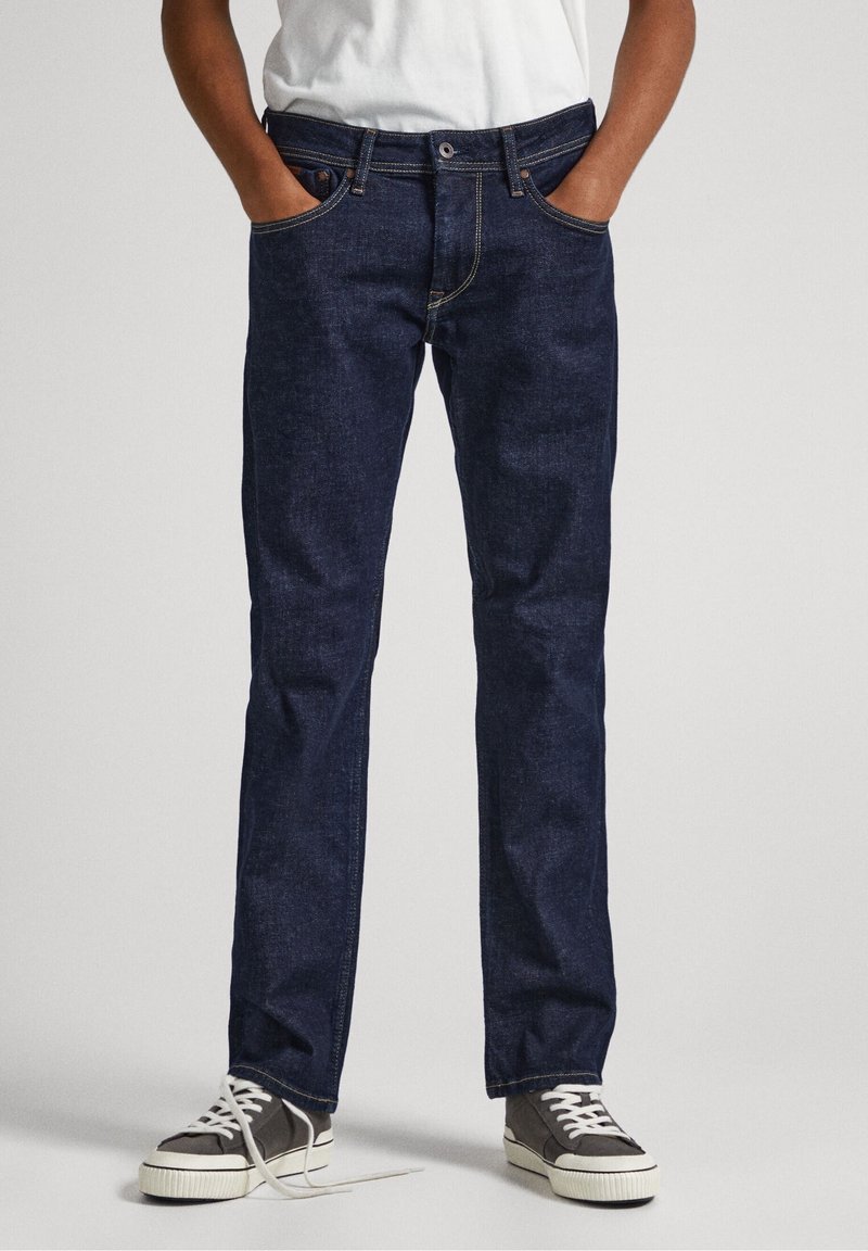 Jeans de denim azul oscuro con un corte recto, bolsillos frontales y costuras en contraste. Combinados con zapatillas grises y blancas.