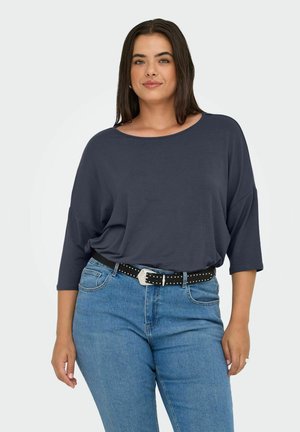 Femme avec des cheveux bruns mi-longs portant un haut ample bleu foncé, une ceinture noire cloutée et un jean bleu, se tenant debout avec une main détendue.