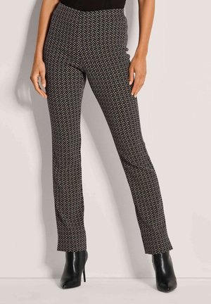 Vrouw in een strakke zwarte en witte geometrische patroon broek met zwarte hoge hakken en enkellaarsjes, staande tegen een effen achtergrond.