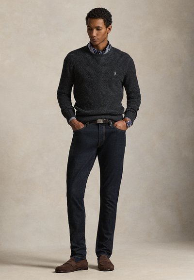 Donkergrijze gebreide trui, donkerblauwe jeans en bruine suède schoenen. Model staat rechtop met handen in de zakken, draagt een overhemd met patroon eronder.