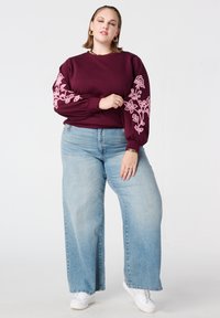Bordeauxrode sweatshirt met roze bloemige borduursels op de opgepofte mouwen, gecombineerd met lichtblauwe wijde jeans en witte sneakers.