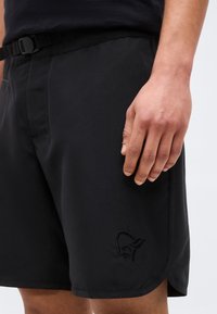 Το χέρι ενός άνδρα ακουμπάει δίπλα σε μαύρο σορτς με ενσωματωμένη ζώνη και διακριτικό λογότυπο ζώου κοντά στο τελείωμα.