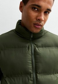 Vadderad grön jacka med hög krage och dragkedjestängning; slät textur; har horisontell sömnad för isolering. Inga synliga hårdvarudetaljer.