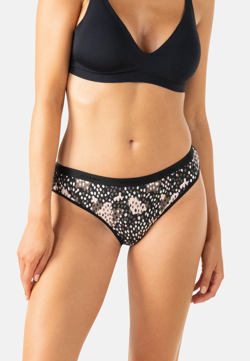 Femme portant un soutien-gorge noir sans coutures et un caleçon hipster à motifs noirs et beiges, sur un fond clair.