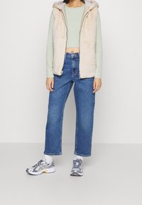 Beżowa kamizelka z sztucznego futra z kapturem, jasnooliwkowa bluzka z długim rękawem, niebieskie jeansy z wysokim stanem oraz szare sportowe buty z białymi akcentami.