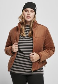 Bruine corduroy jas met een hoge kraag en ribbeltextuur, gecombineerd met een grijs gestreept shirt eronder. Zwarte gebreide beanie als accessoire.