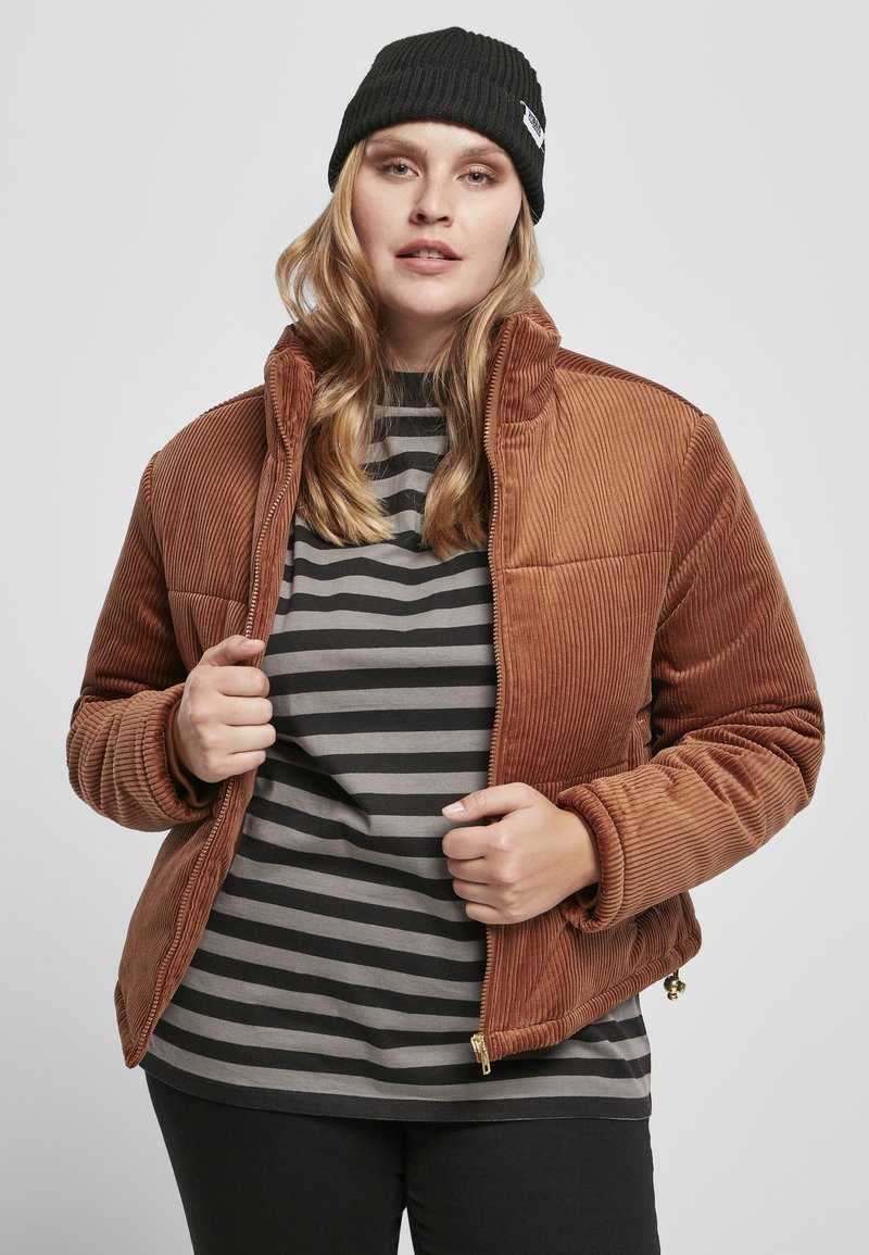 Bruine corduroy jas met een hoge kraag en ribbeltextuur, gecombineerd met een grijs gestreept shirt eronder. Zwarte gebreide beanie als accessoire.