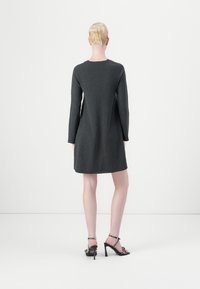 Femme aux cheveux blonds en chignon, portant une robe gris anthracite à manches longues, longueur genou, et des sandales noires à talons hauts avec des brides à la cheville.