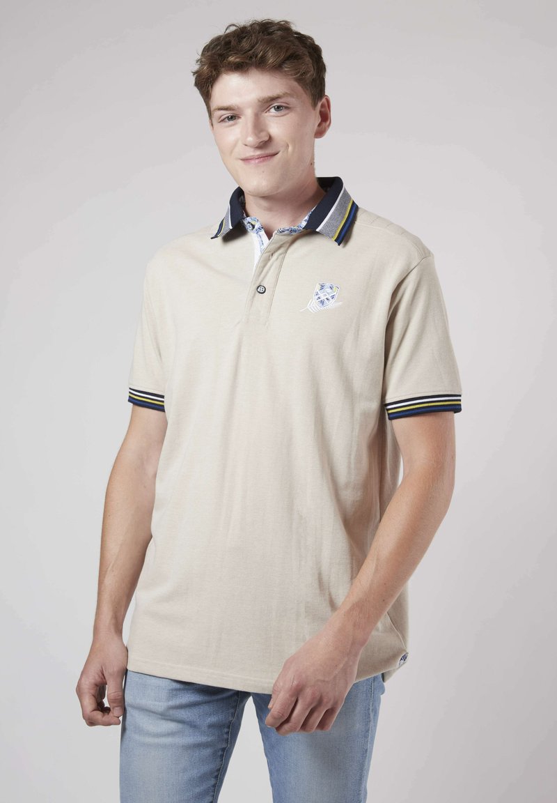Polo beige avec un col marine, des accents rayés et un logo sur la poitrine. Manches courtes, tissu texturé et patte de boutonnage.