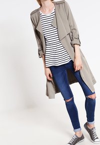 Trinchera gris claro con frente abierto, combinada con una blusa peplum a rayas blancas y negras y jeans skinny azules desgastados. Zapatillas grises completan el look.