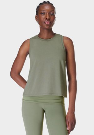 Vrouw met gevlochten haar draagt een olijfgroene mouwloze top en bijpassende legging, staand met één hand achter haar rug.