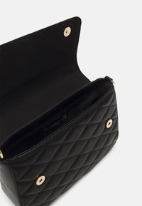 Sac à main en cuir matelassé noir avec une fermeture à bouton-pression doré. À l'intérieur, il est doté d'une doublure noire lisse et d'une étiquette. Forme rectangulaire.