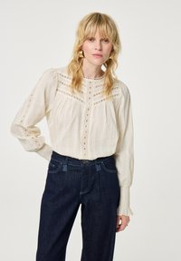 Langere crème blouse met een textuurpatroon, ronde halslijn, knoopdetails en gerimpelde manchetten, gecombineerd met hoge taille donkere denim broek.