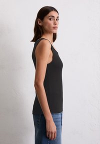 Femme aux cheveux foncés à mi-longueur portant un haut sans manches noir et un jean bleu, se tenant contre un mur blanc texturé, de profil.
