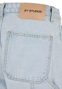 Jeans din denim de un albastru deschis, dotate cu un buzunar mare la spate, cusături contrastante în portocaliu și un etichetă din piele pal, embosată cu textul "2Y STUDIOS."