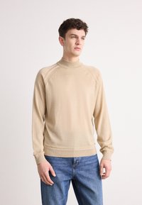 Beige høyhalset genser med lange ermer, ribbede mansjetter og en jevn strikket tekstur. Brukt med blå jeans; enkel bakgrunn.